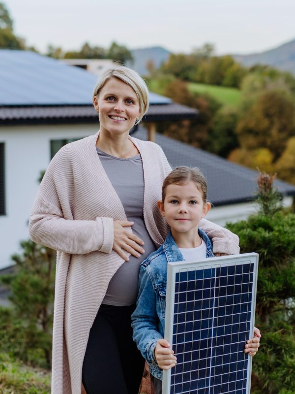 Mother with her little daughter near their house with solar panels. Alternative energy, saving